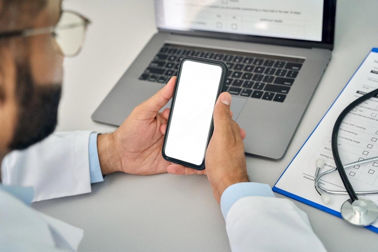 Medical professional in a white coat holding a smartphone with a blank screen next to a laptop and a clinical checklist